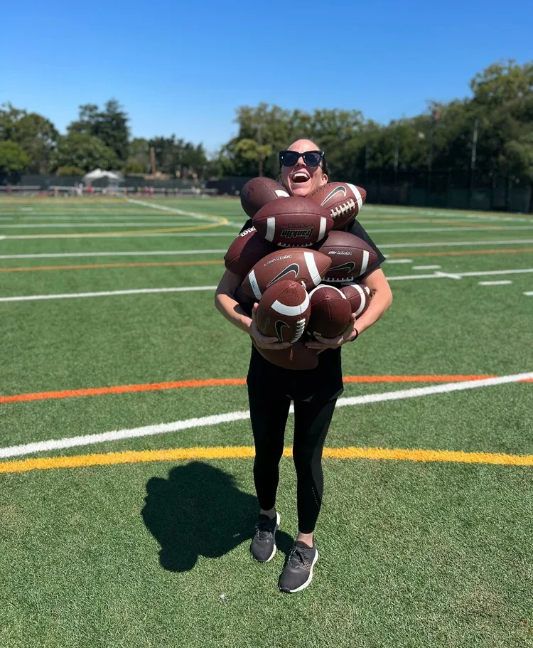 A smiling coach on a turf field carrying an armful of footballs