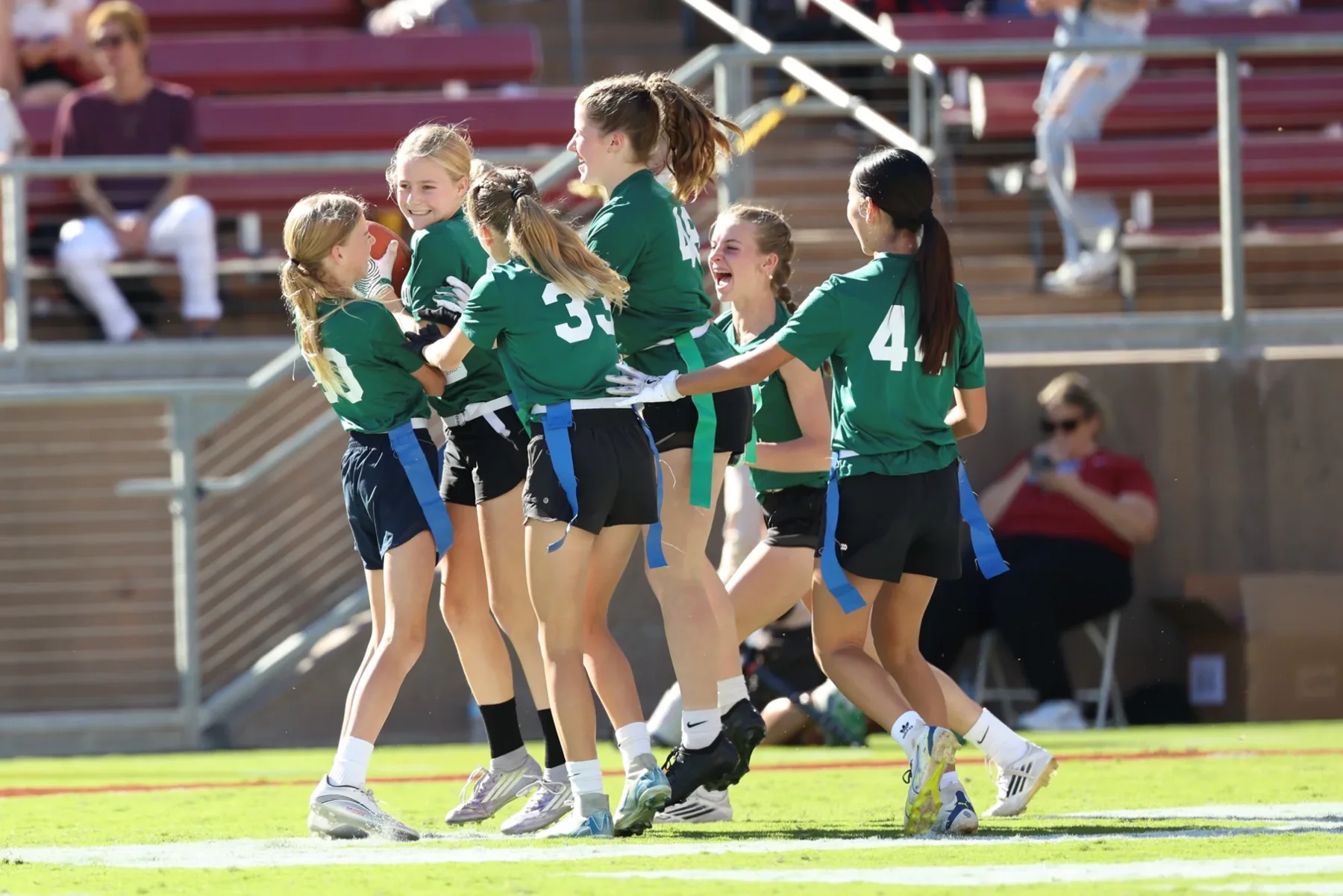Girls in green jerseys and flag belts celebrating together on the field after a play